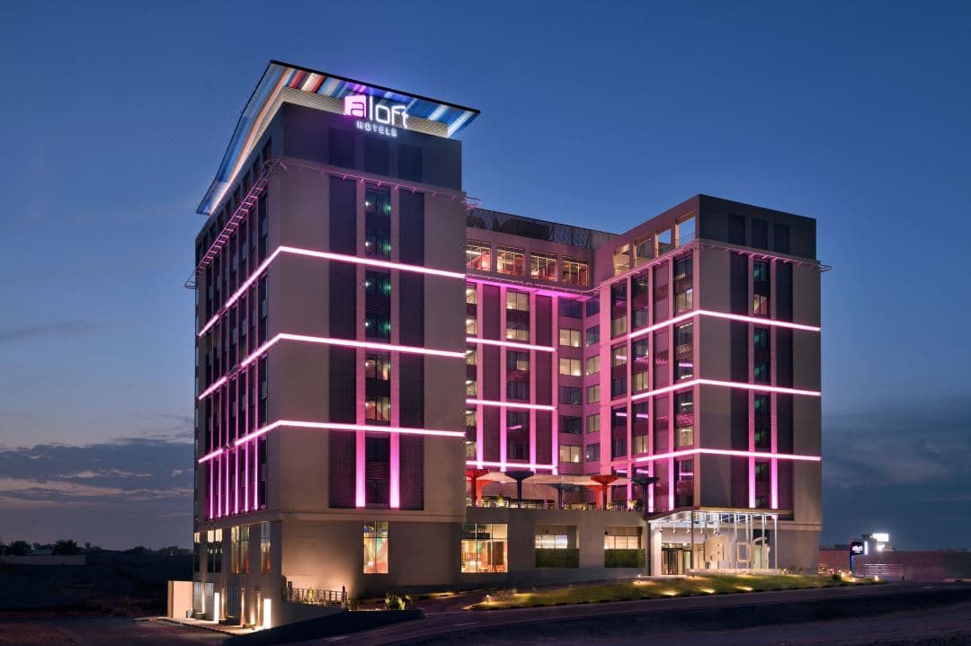 Rooftop infinity pool at Aloft Muscat at twilight with illuminated city skyline and guests enjoying cocktails.