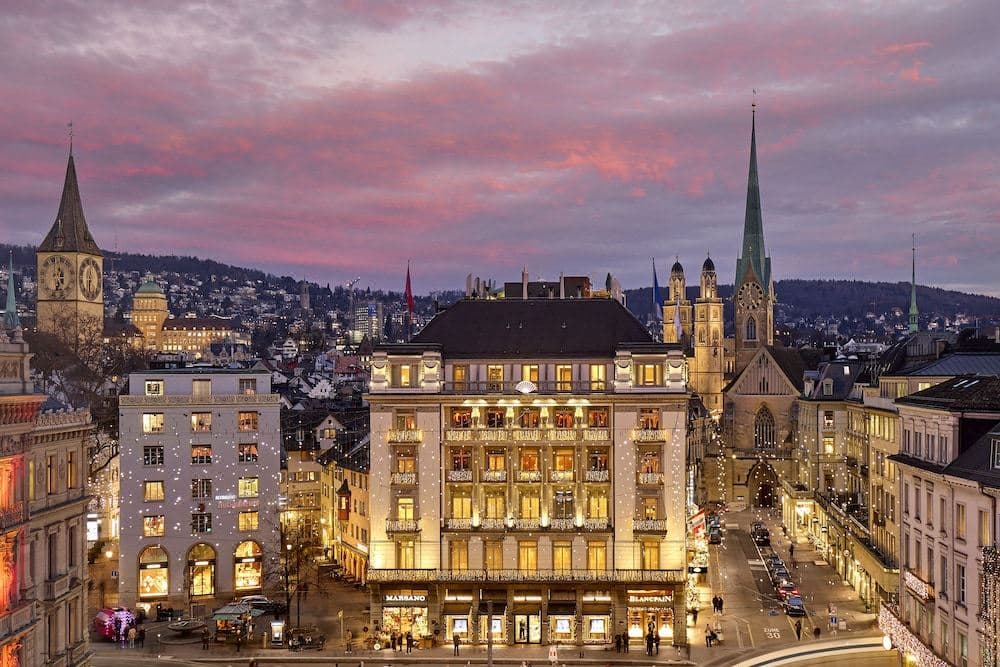 A panoramic view of the 1838 Rooftop Bar at Mandarin Oriental Savoy Zurich with guests enjoying cocktails against a backdrop of the city and distant Alps.