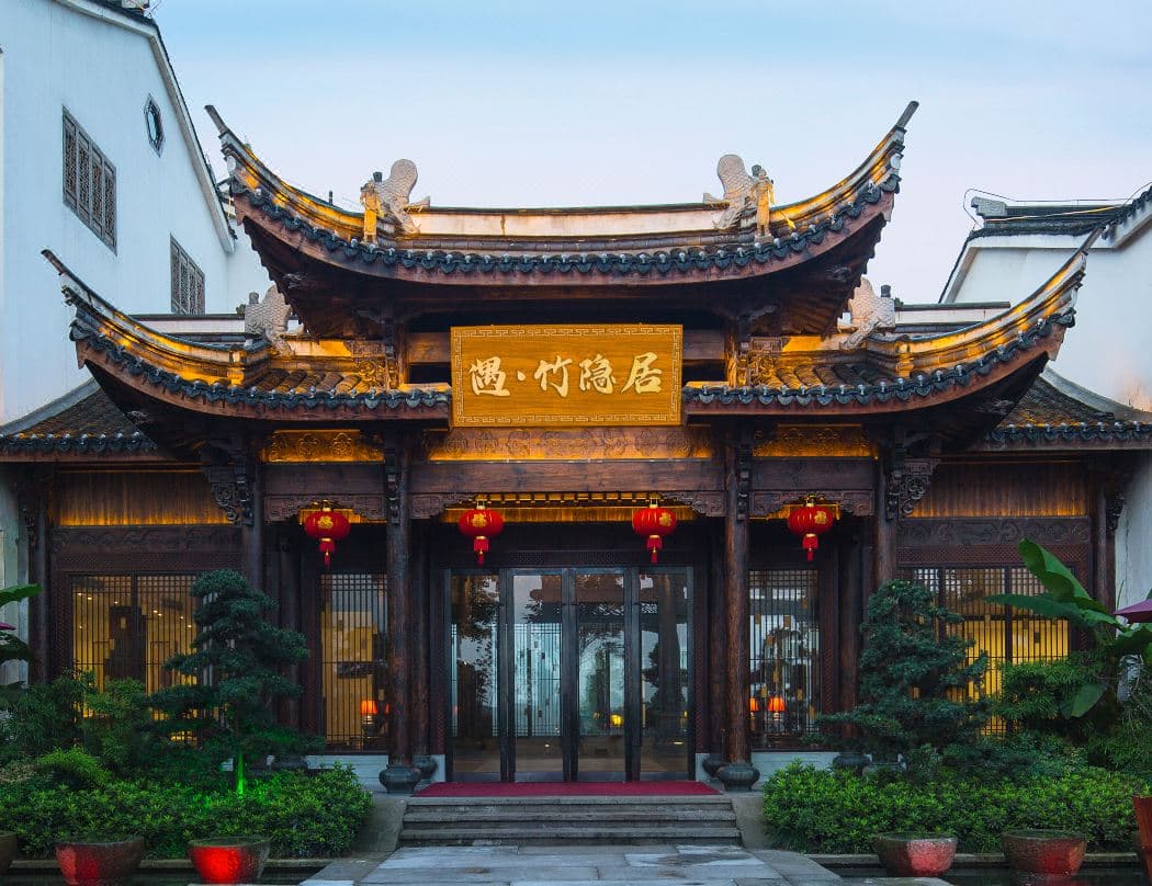 A serene courtyard at Liyang Yuzhu Reclusive Hotel with wooden architecture, glowing lanterns at twilight, and a backdrop of dense bamboo forest.