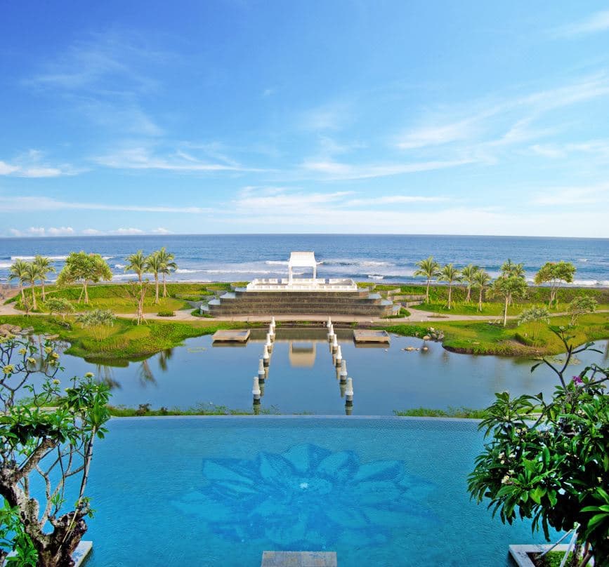 The jade-green infinity pool of Rumah Luwih Bali contrasting against the black volcanic sand of Lebih Beach, framed by white colonial architecture.