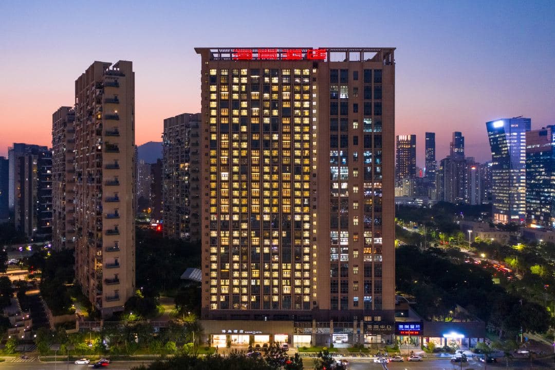 A spacious hotel suite with floor-to-ceiling windows overlooking the Shenzhen Bay bridge and the Spring Cocoon stadium during twilight.