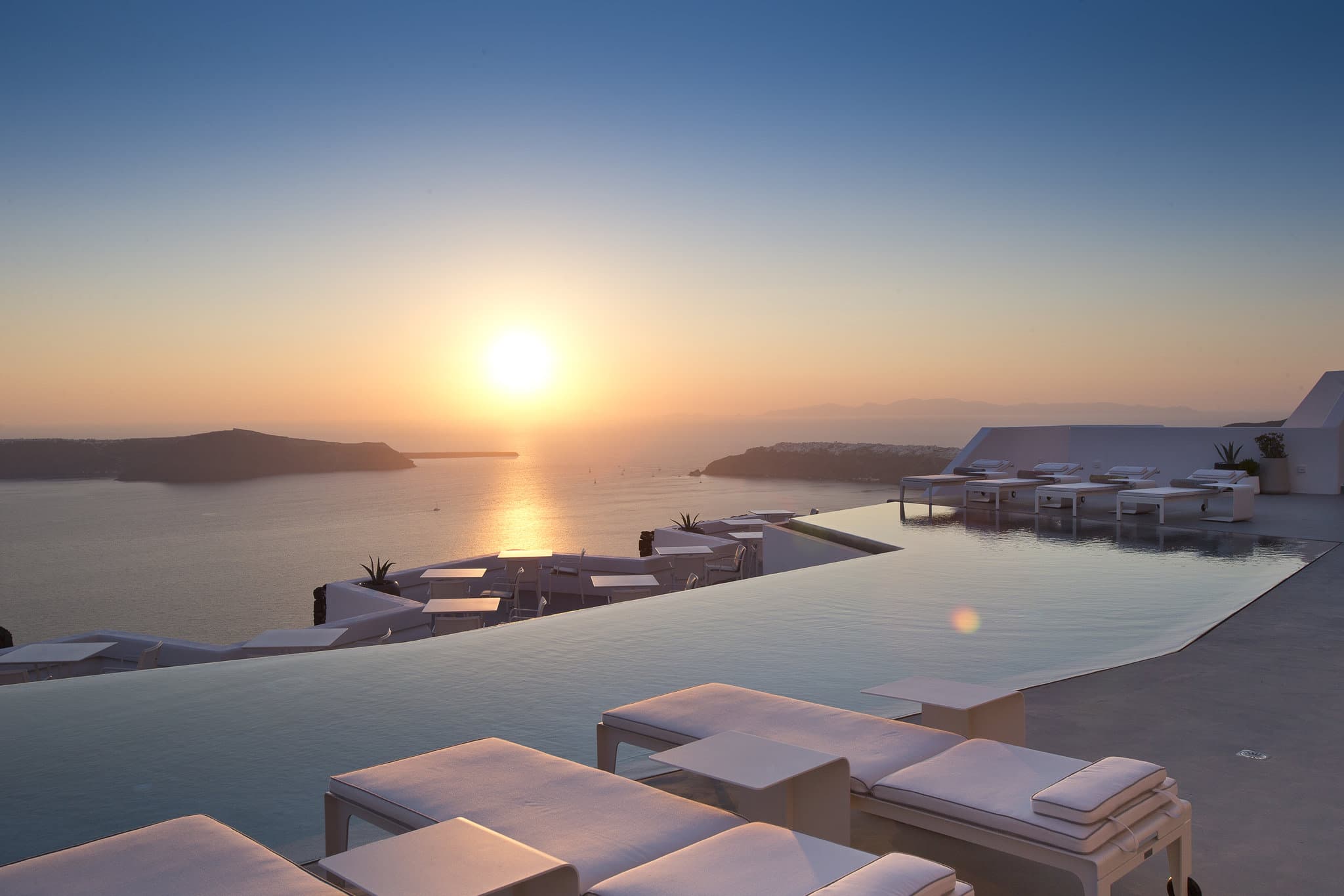A woman in a white dress standing by the 22-meter infinity pool at Grace Hotel Santorini, looking out at Skaros Rock and the Aegean Sea sunset.