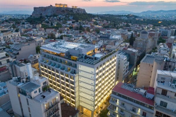A dramatic dusk view from the Electra Metropolis rooftop restaurant, with guests dining in soft lighting directly facing the illuminated Parthenon and Acropolis hill.