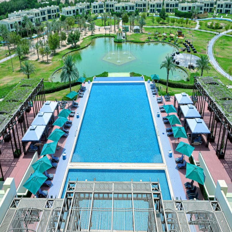 A wooden arched bridge spanning the winding lazy river at Retaj Salwa Resort, surrounded by palm trees.