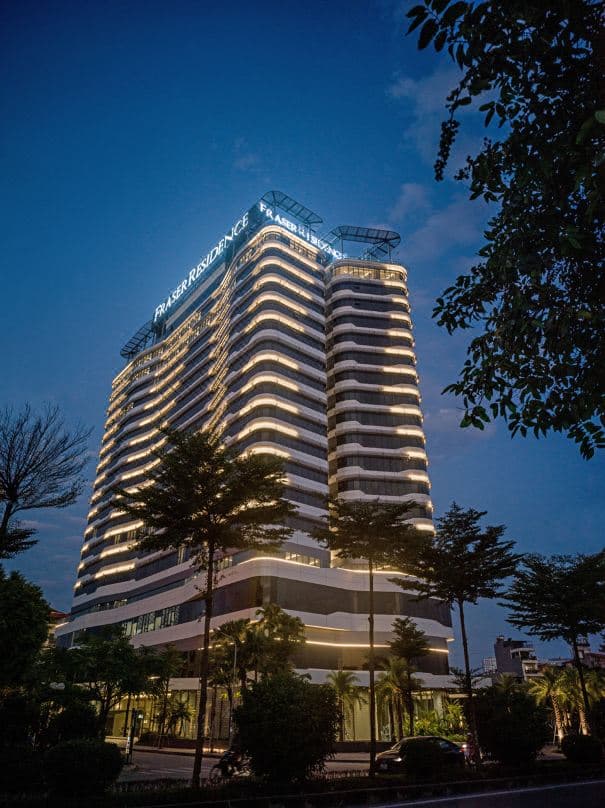 A sleek, modern residence facade reflecting into a serene outdoor pool, with Hoa Binh Park greenery in the background.