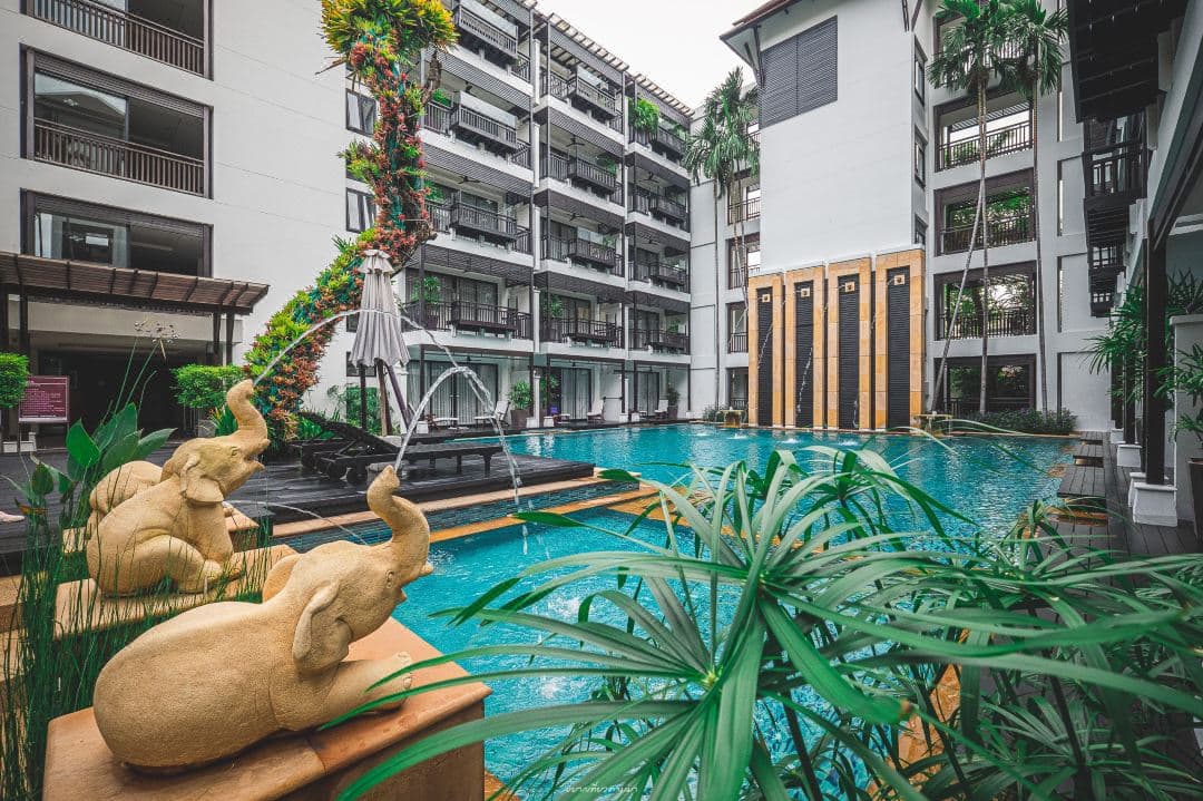 A twilight view of the RarinJinda swimming pool reflecting the illuminated 140-year-old teak house, with frangipani trees framing the scene.