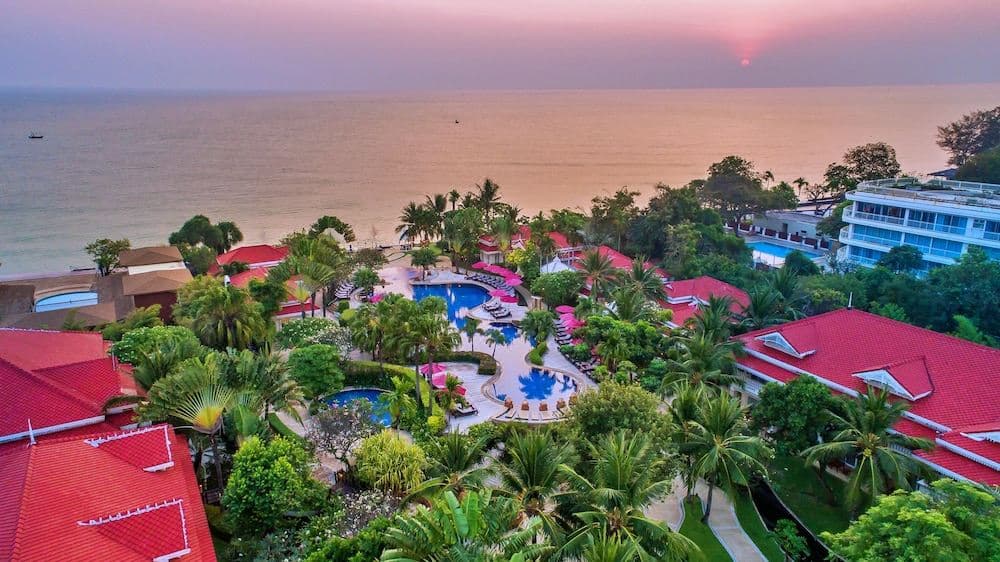 White colonial-style Thai buildings with red roofs reflecting in a large, curving blue lagoon pool surrounded by palm trees.