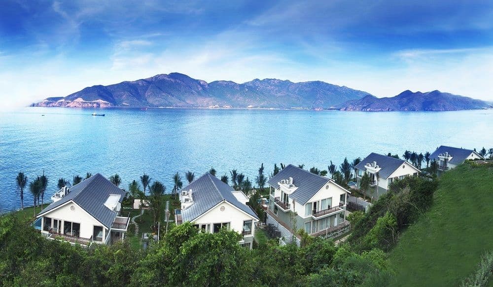 A panoramic view of the MerPerle Mud Bath infinity pool at Hon Tam Resort, overlooking Nha Trang Bay with lush green hills in the background.