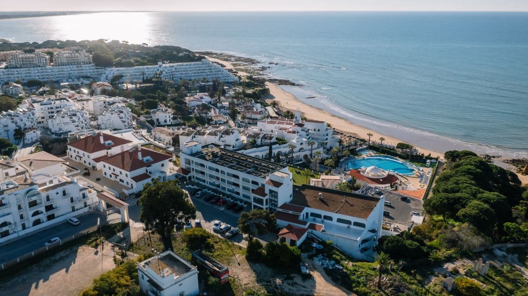 Grand Muthu Oura View Beach Clubの巨大な楕円形インフィニティプールが、青い大西洋と一体化し、レトロな白い建築とヤシの木に囲まれている様子。