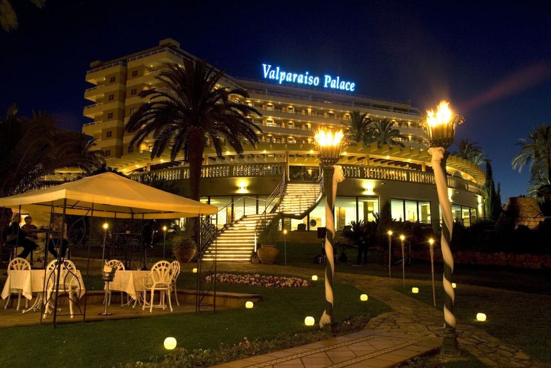 A wide shot of the GPRO Valparaiso Palace outdoor saltwater pools surrounded by lush gardens, with the city of Palma and the sea visible in the background.