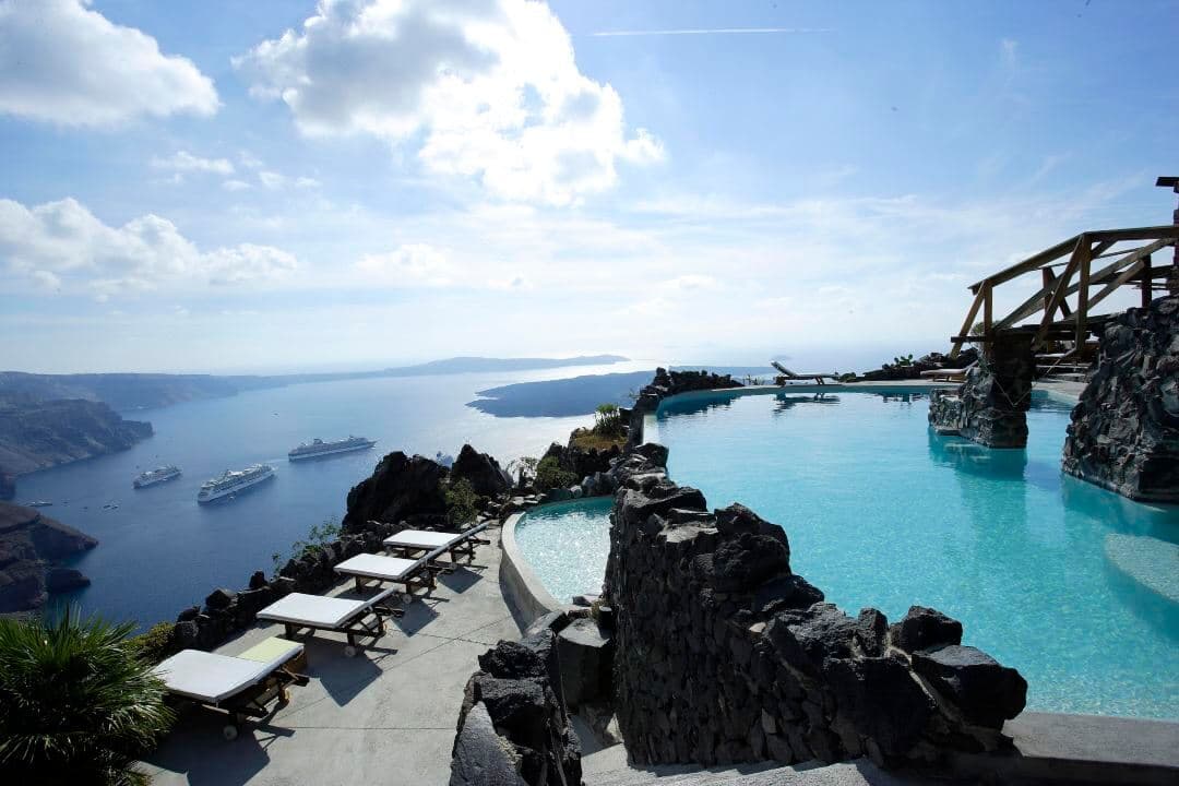 Honeymoon Petra Villas infinity pool carved into volcanic rock overlooking Skaros Rock and the Santorini caldera at sunset.