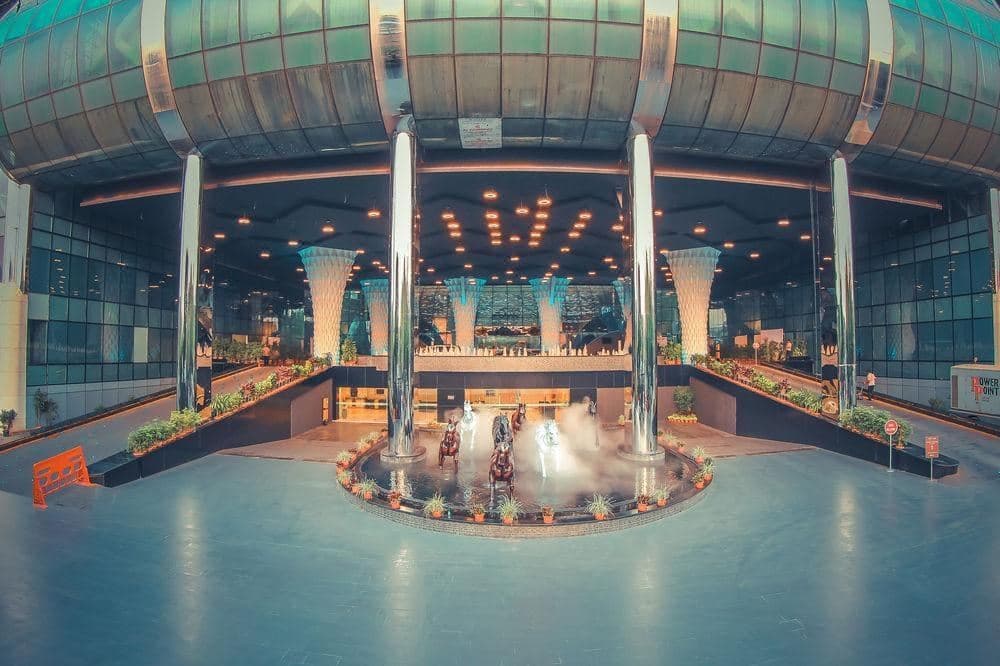 Glass elevator rising through a massive indoor tropical atrium with a blue lagoon and illuminated dome roof at Hotel Sahara Star.