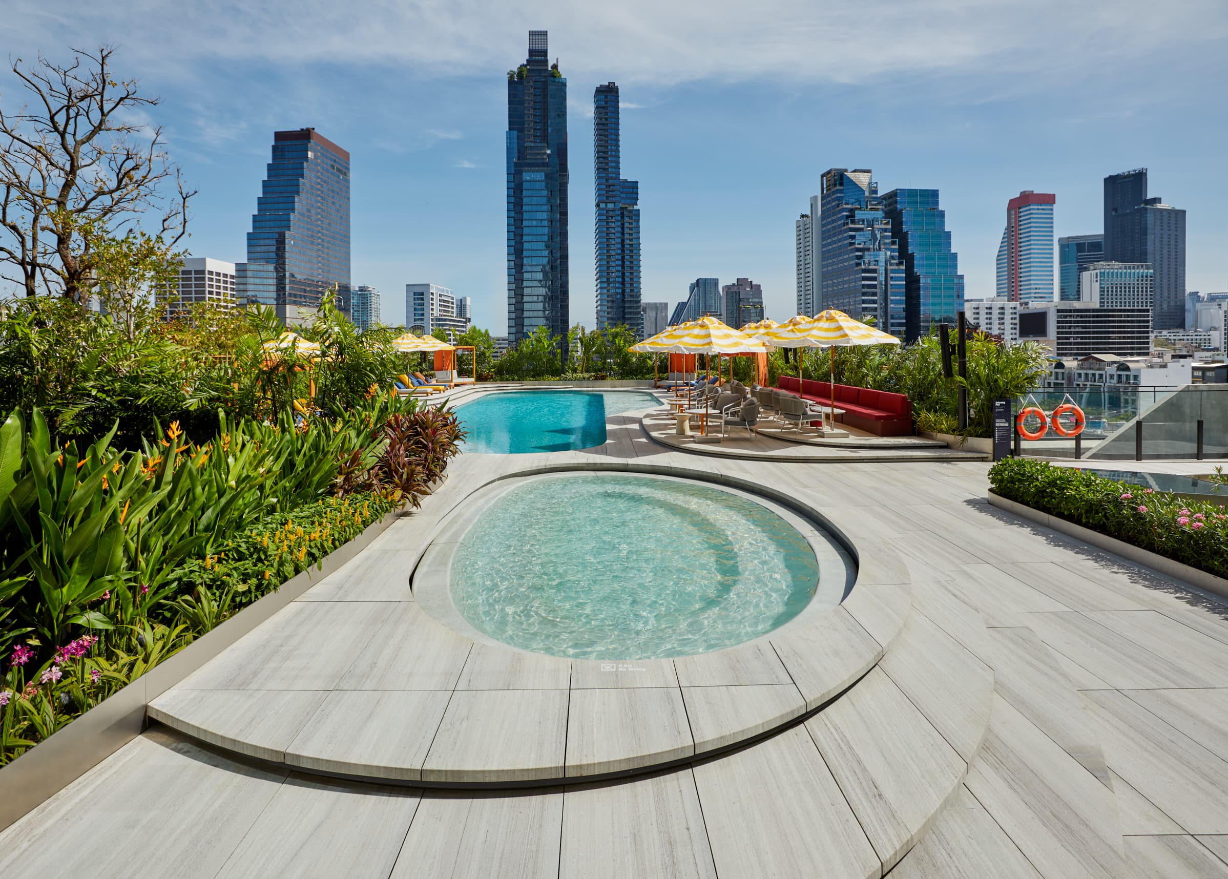 A low-angle shot of The Standard Bangkok's terrace pool area featuring signature mustard-yellow striped umbrellas against the pixelated backdrop of the Mahanakhon building.