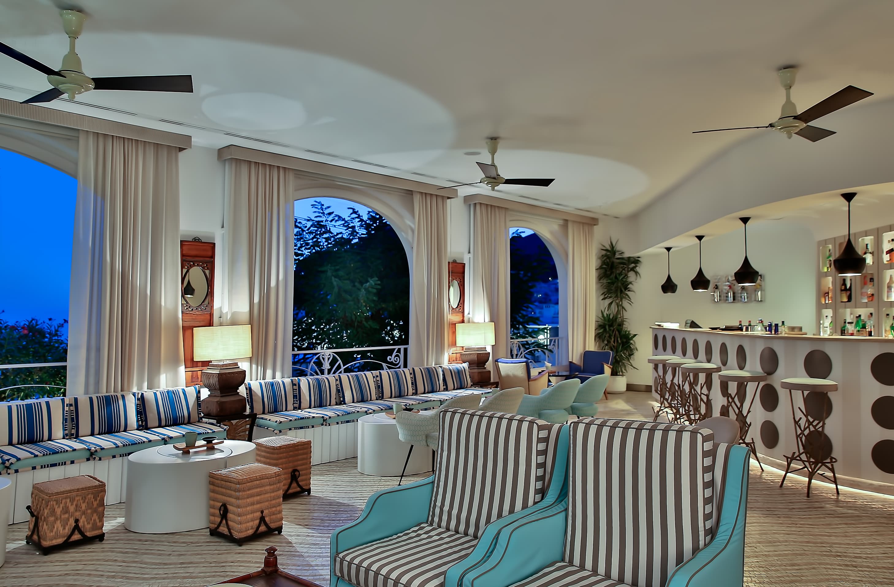 A vibrant hotel terrace in Capri featuring a white grand piano, a wall of Panama hats, and colorful 1950s-style furniture overlooking the sea.