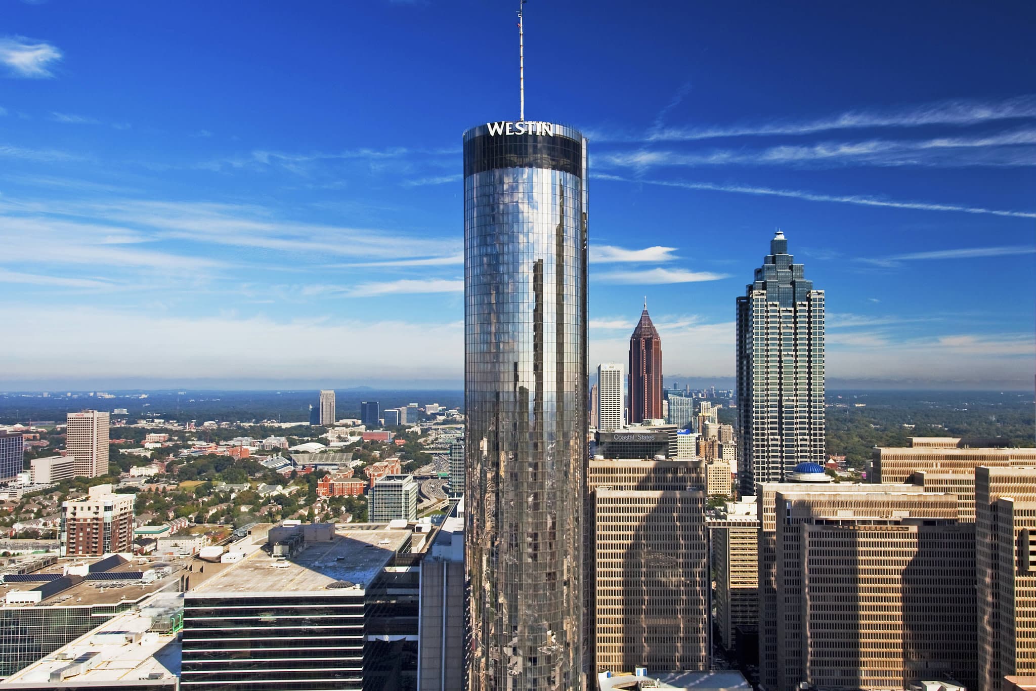 A low-angle shot of The Westin Peachtree Plaza's cylindrical glass tower reflecting the blue Atlanta sky, with its signature external glass elevator shaft visible.