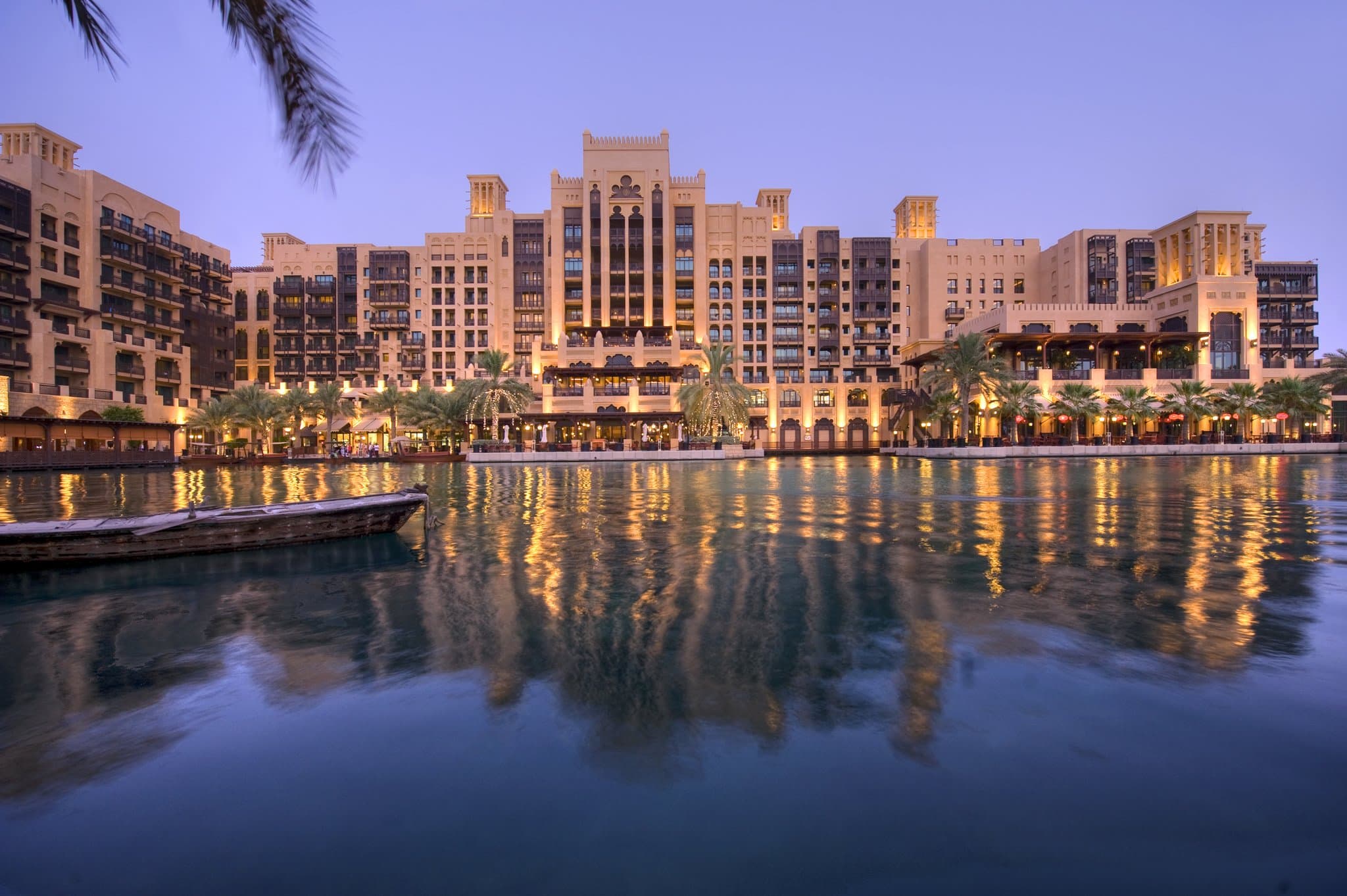 A traditional abra boat gliding through the turquoise canals of Madinat Jumeirah with the wind-tower architecture of Mina A'Salam and the Burj Al Arab in the background.