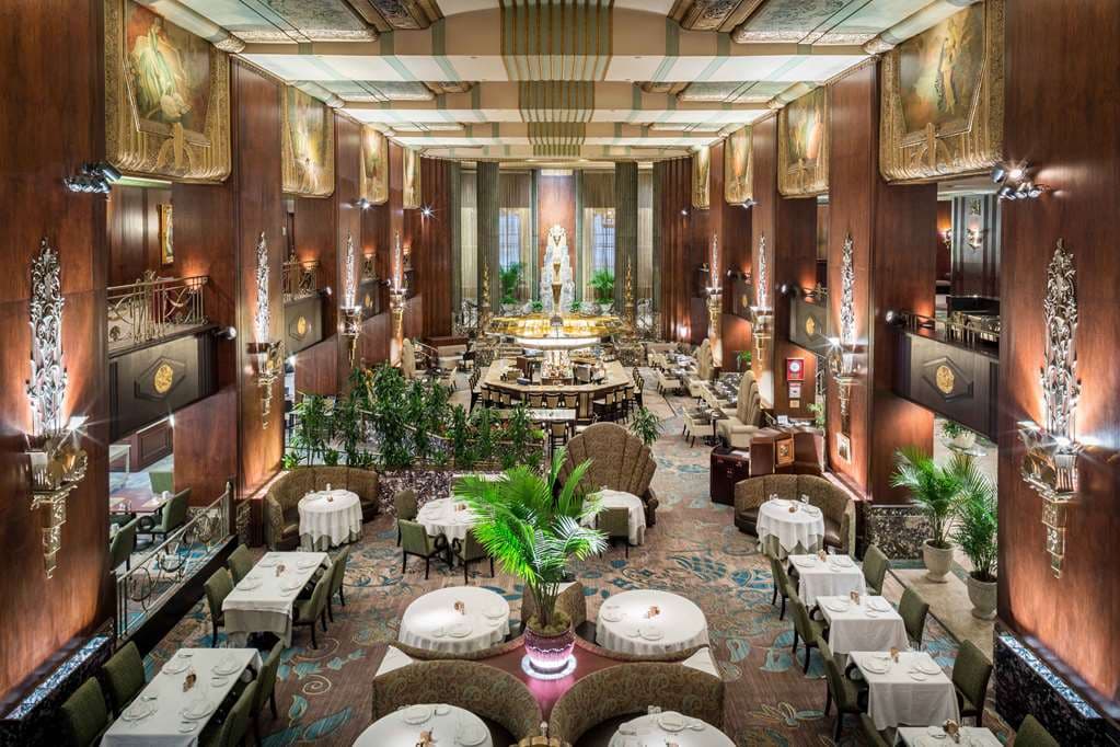 Wide angle shot of the Art Deco Palm Court with Brazilian rosewood paneling, ceiling frescoes, and the Rookwood fountain.