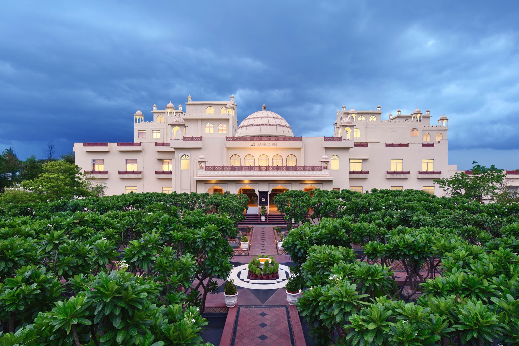 Symmetrical water channels of the Chaupad arrival court at Le Méridien Jaipur, flanked by cream sandstone arches and frangipani trees.