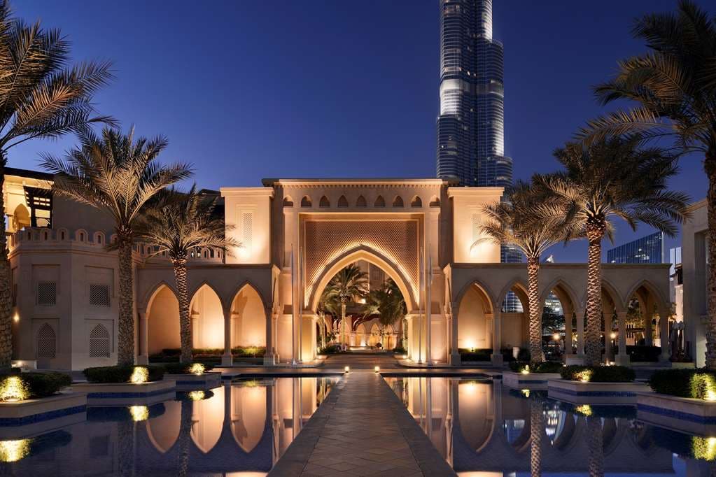 A low-angle night shot of the Palace Downtown's illuminated archway framing the Burj Khalifa, with the Dubai Fountain in the foreground.