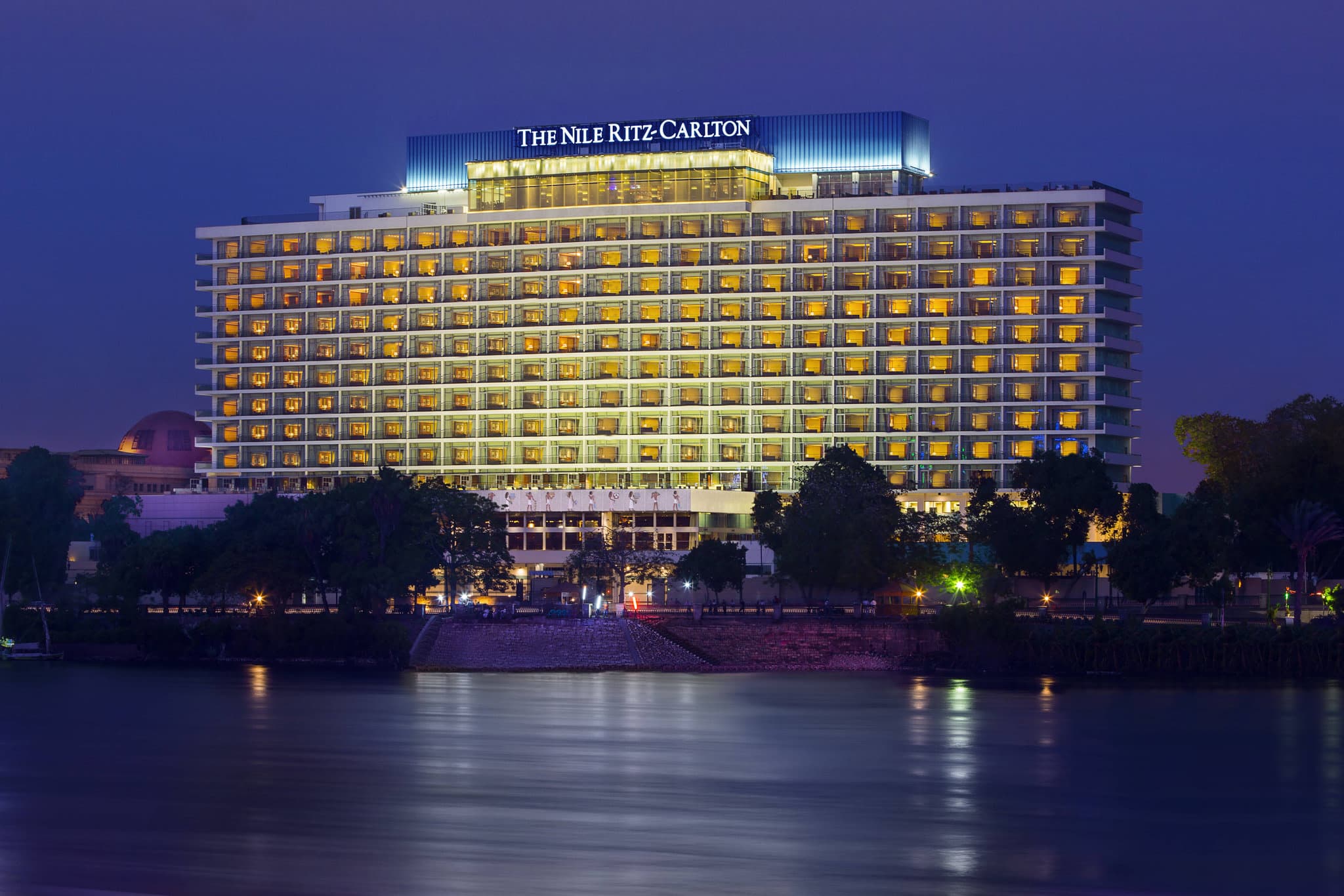 The Nile Ritz-Carlton rooftop pool deck at twilight, featuring private cabanas and a view of the Cairo Tower illuminated in the distance.