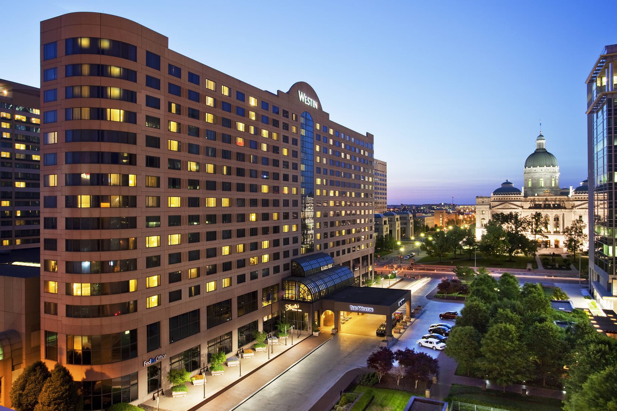A low-angle shot of The Westin Indianapolis facade at twilight, capturing the modern glass exterior and the illuminated skywalk connecting to the city center.