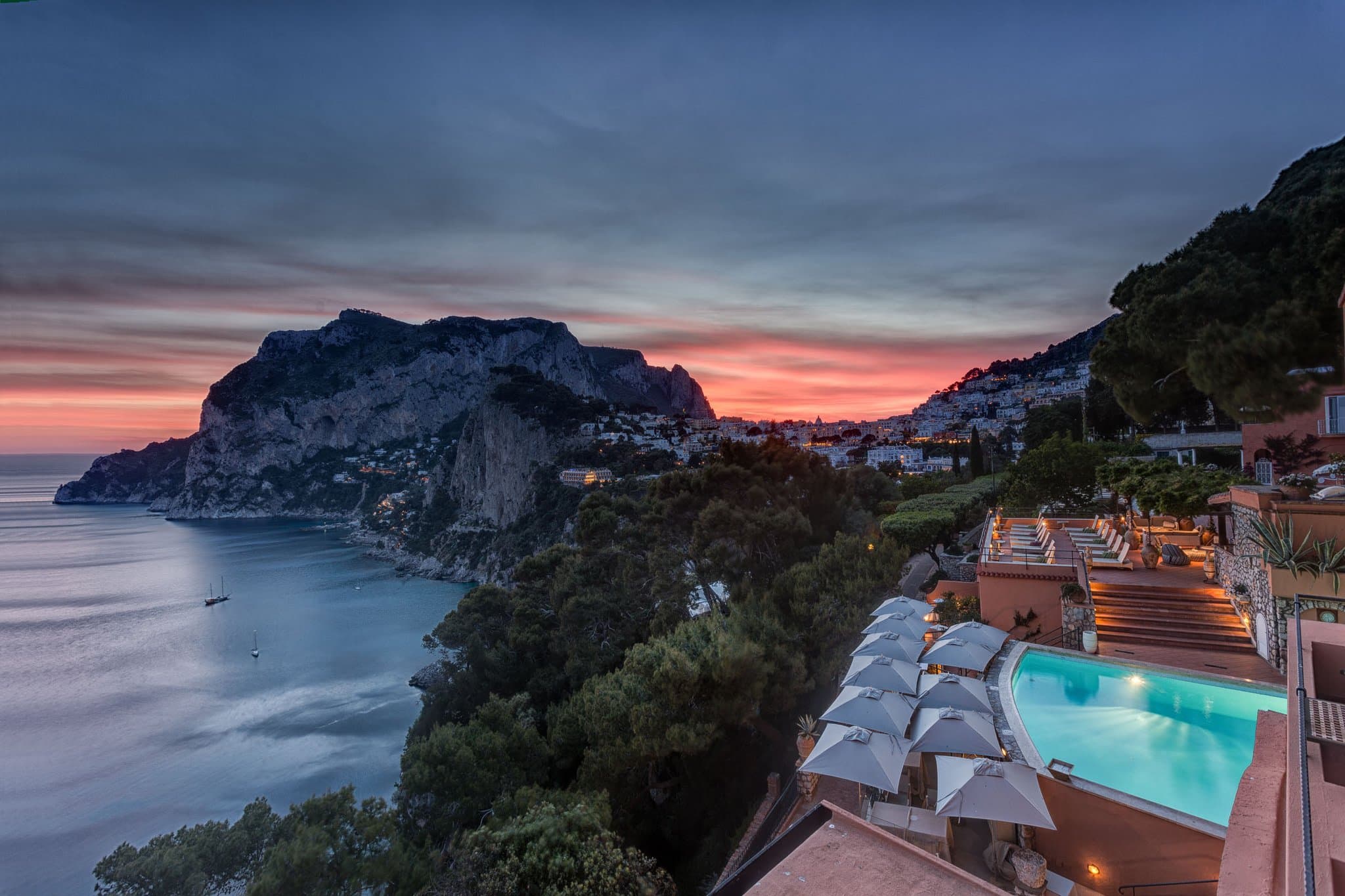 A dramatic sunset view from the Hotel Punta Tragara terrace overlooking the Faraglioni rocks, featuring the Monzù Gin Club setup.