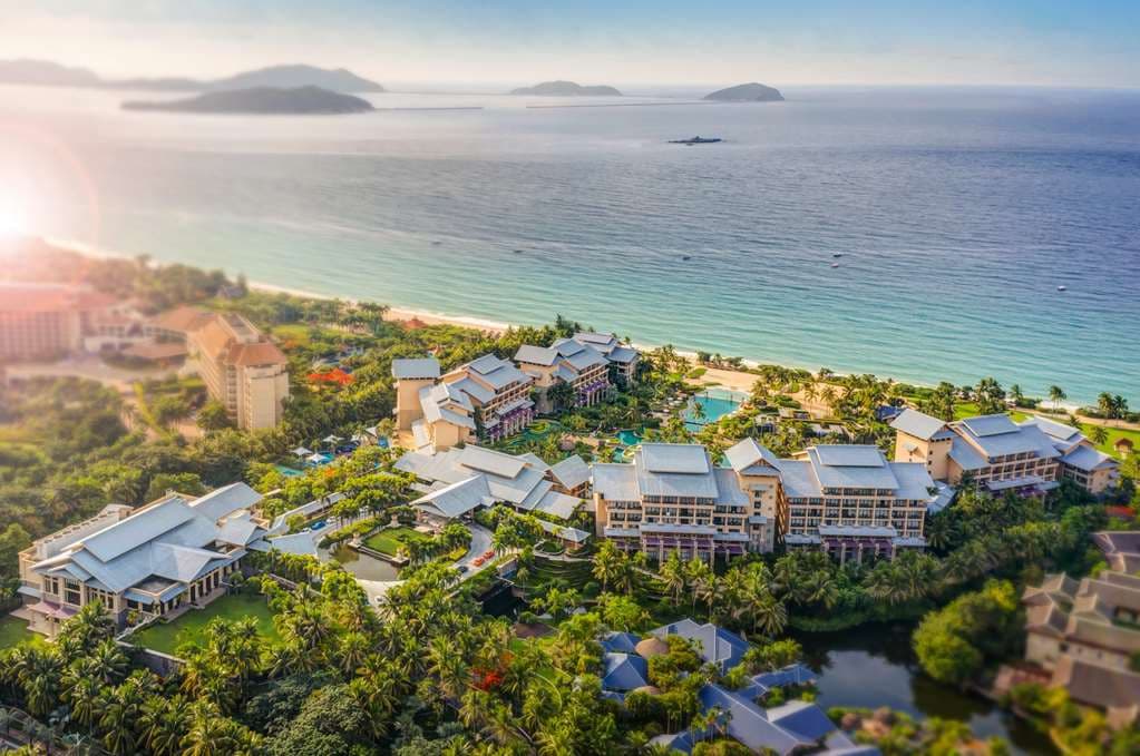 A sprawling lagoon pool at Hilton Sanya Yalong Bay surrounded by mature palm trees and open-air teak pavilions under a blue sky.