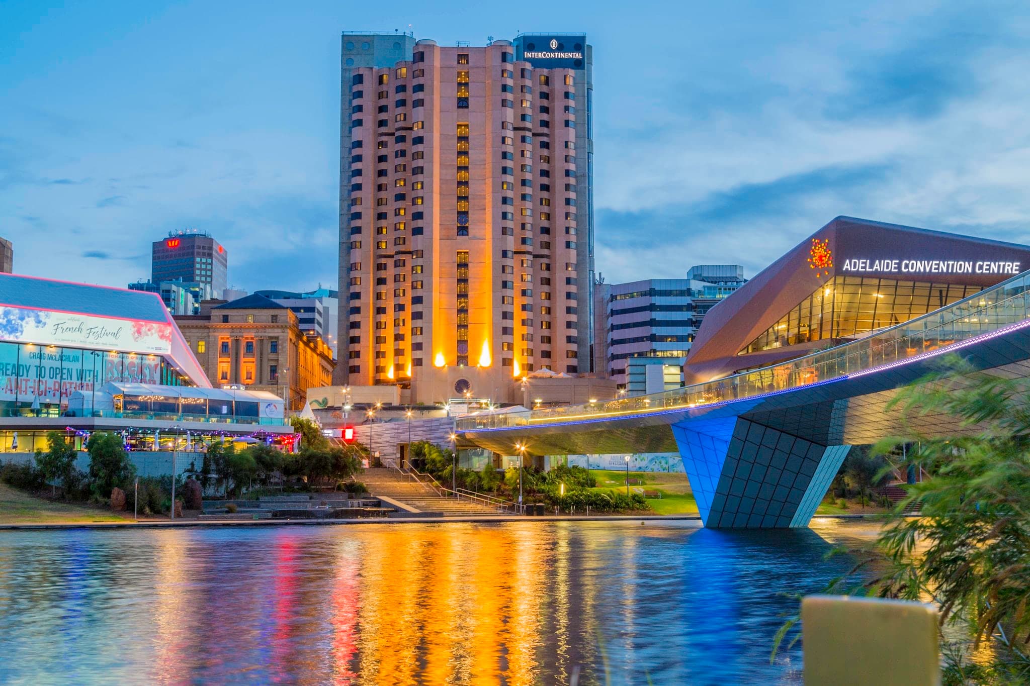 A high-angle view of the InterContinental Adelaide's outdoor pool deck overlooking the River Torrens and the Adelaide Oval footbridge at dusk.