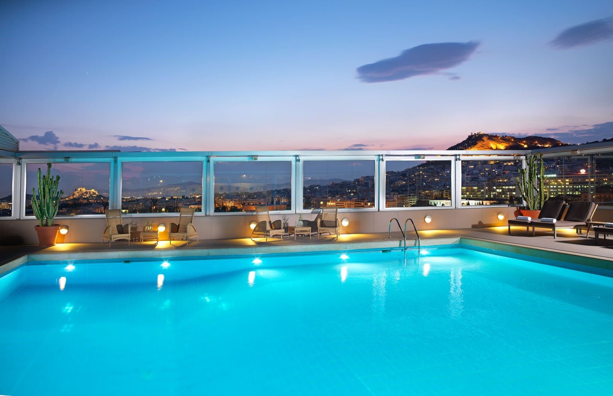 Sunset over the Divani Caravel rooftop pool, known as Thea, featuring guests with cocktails and a lit Acropolis in the background.