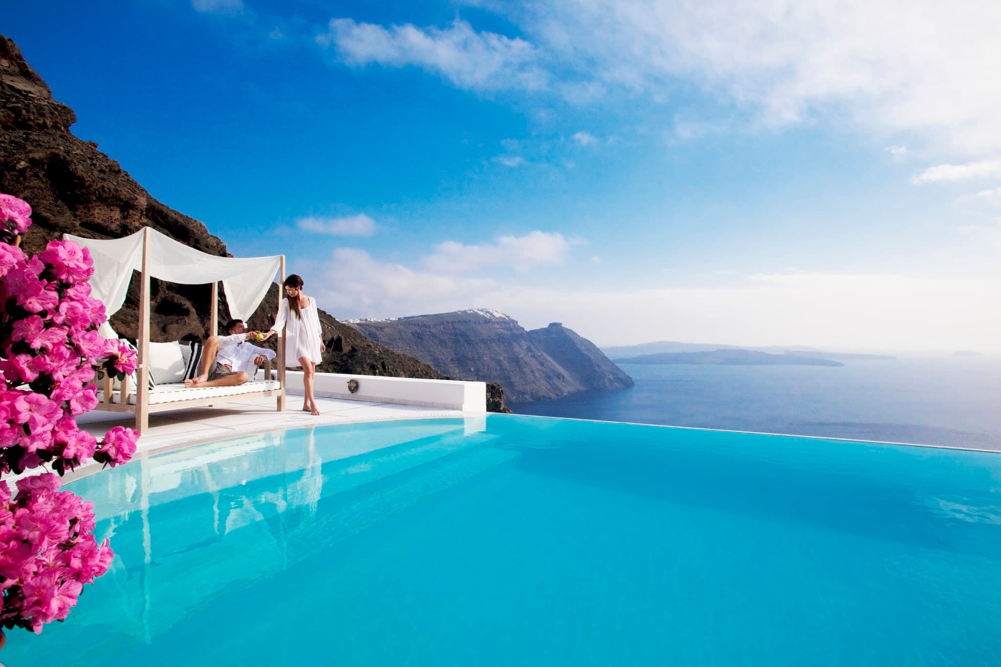 San Antonio hotel infinity pool suspended 100m above the Santorini caldera, framed by natural red volcanic rock.