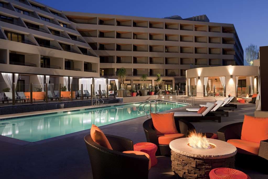 A dusk view of the Hyatt Palm Springs HooDoo Patio featuring fire pits and guests dining, with the illuminated geometric hotel facade and San Jacinto mountains in the background.