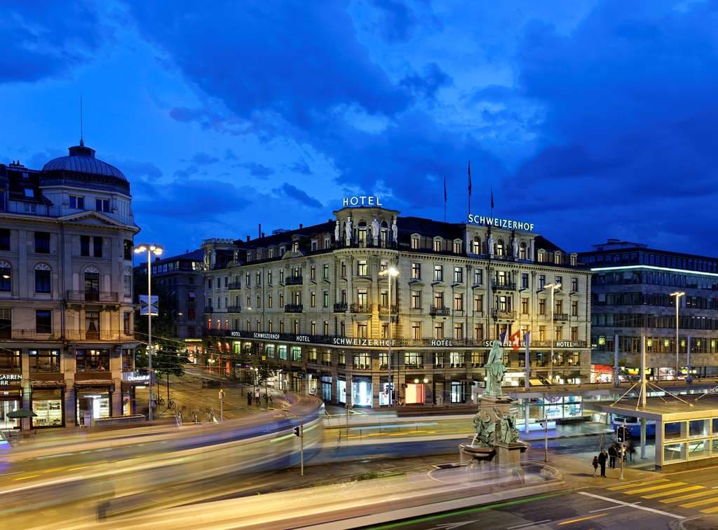 A butler adjusting fresh flowers in a sunlit Hotel Schweizerhof suite overlooking Zurich Main Station.