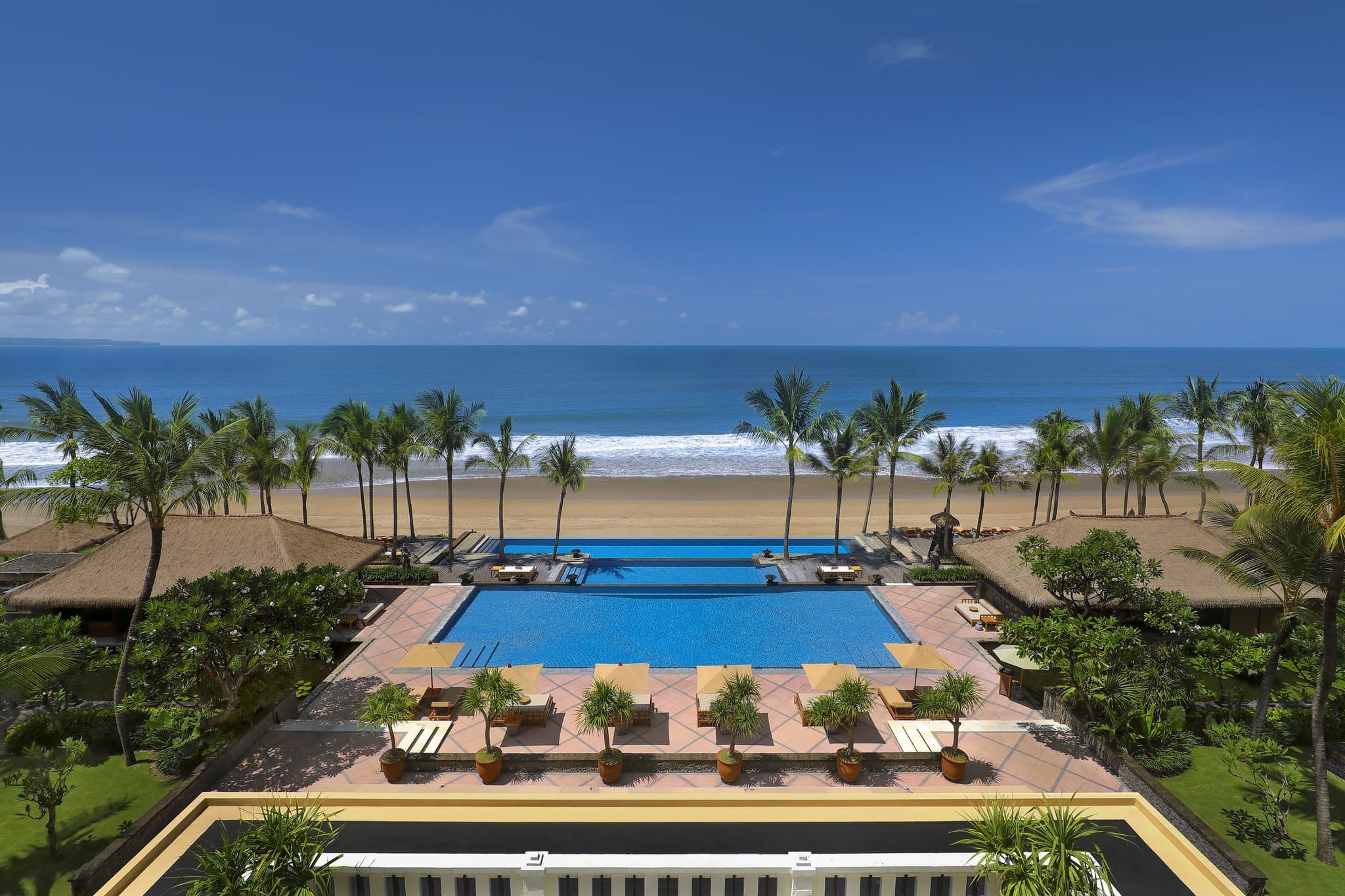 The Legian Seminyak's three-tiered infinity pool cascading towards the Indian Ocean at sunset, framed by frangipani trees.