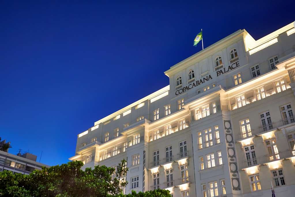 The illuminated white facade of the Copacabana Palace at twilight, reflecting onto the semi-Olympic pool with palm trees.