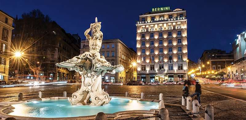 A low-angle view of the Sina Bernini Bristol's exterior featuring the Triton Fountain in the foreground under a dusk sky.