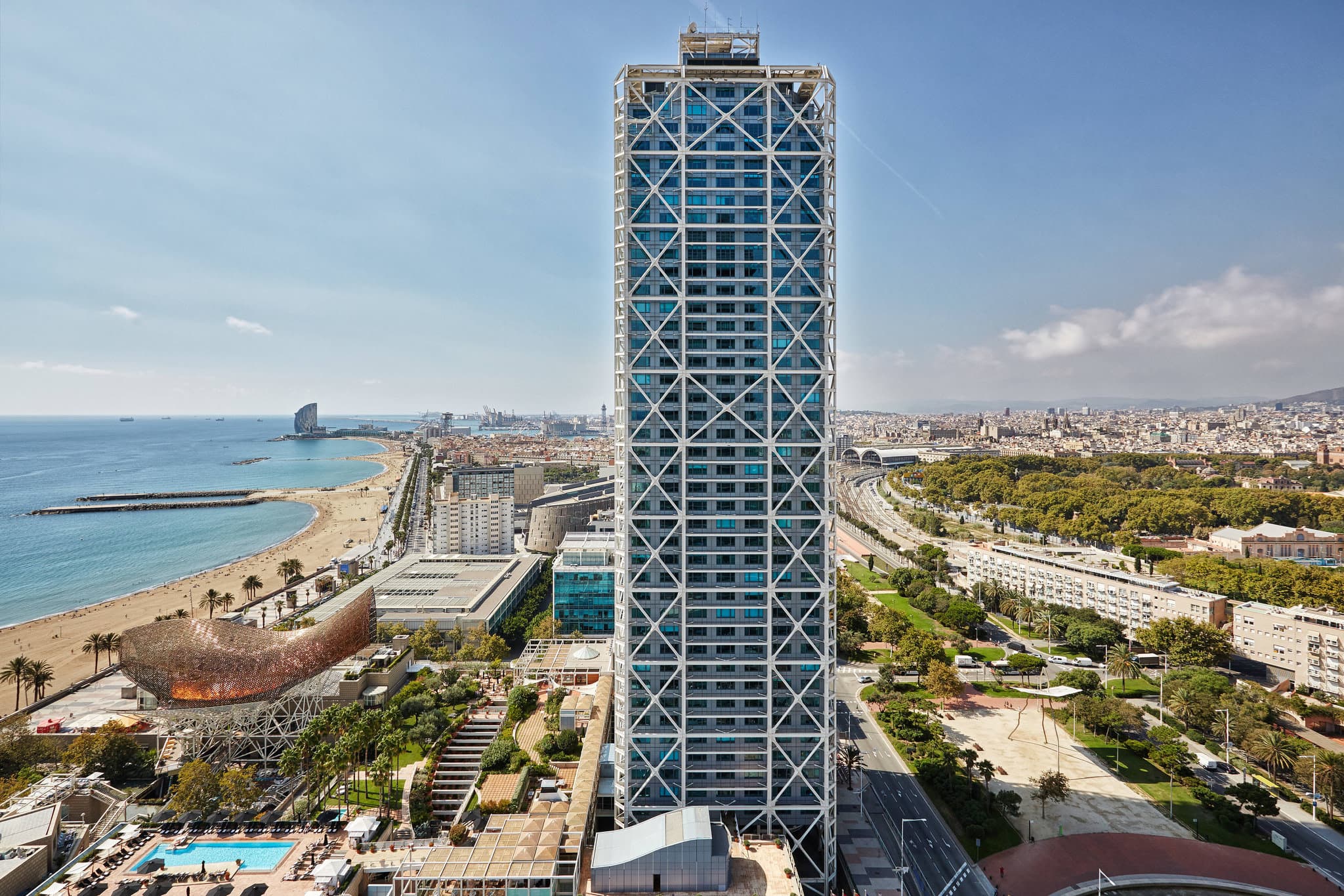 The exposed white steel beams of Hotel Arts Barcelona framing a view of Frank Gehry's golden 'Peix' sculpture against the blue Mediterranean Sea.