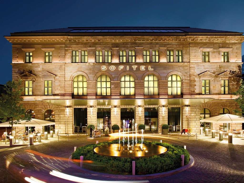 The illuminated spiral-shaped indoor lagoon pool at Sofitel Munich Bayerpost, surrounded by stone columns and lounge chairs.