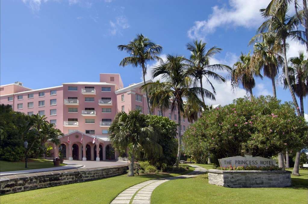 The iconic pink facade of the Hamilton Princess Bermuda viewed from the marina, featuring the KAWS 'At This Time' statue.