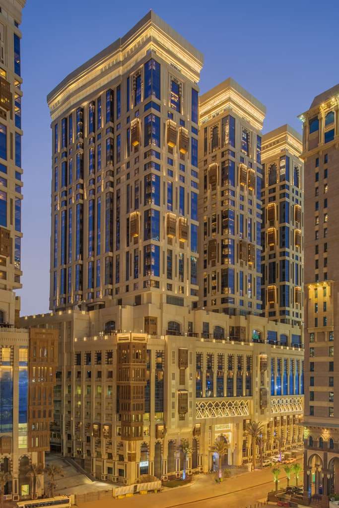 A dramatic low-angle view of the Hyatt Regency Makkah's crystal chandeliers in the lobby, contrasting with the bustle of pilgrims.