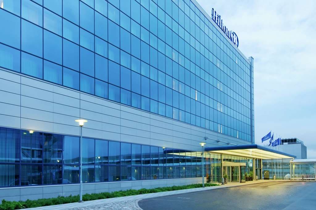 A sleek, glass-enclosed walkway connecting Hilton Helsinki Airport to the terminal, illuminated against a twilight Nordic sky.