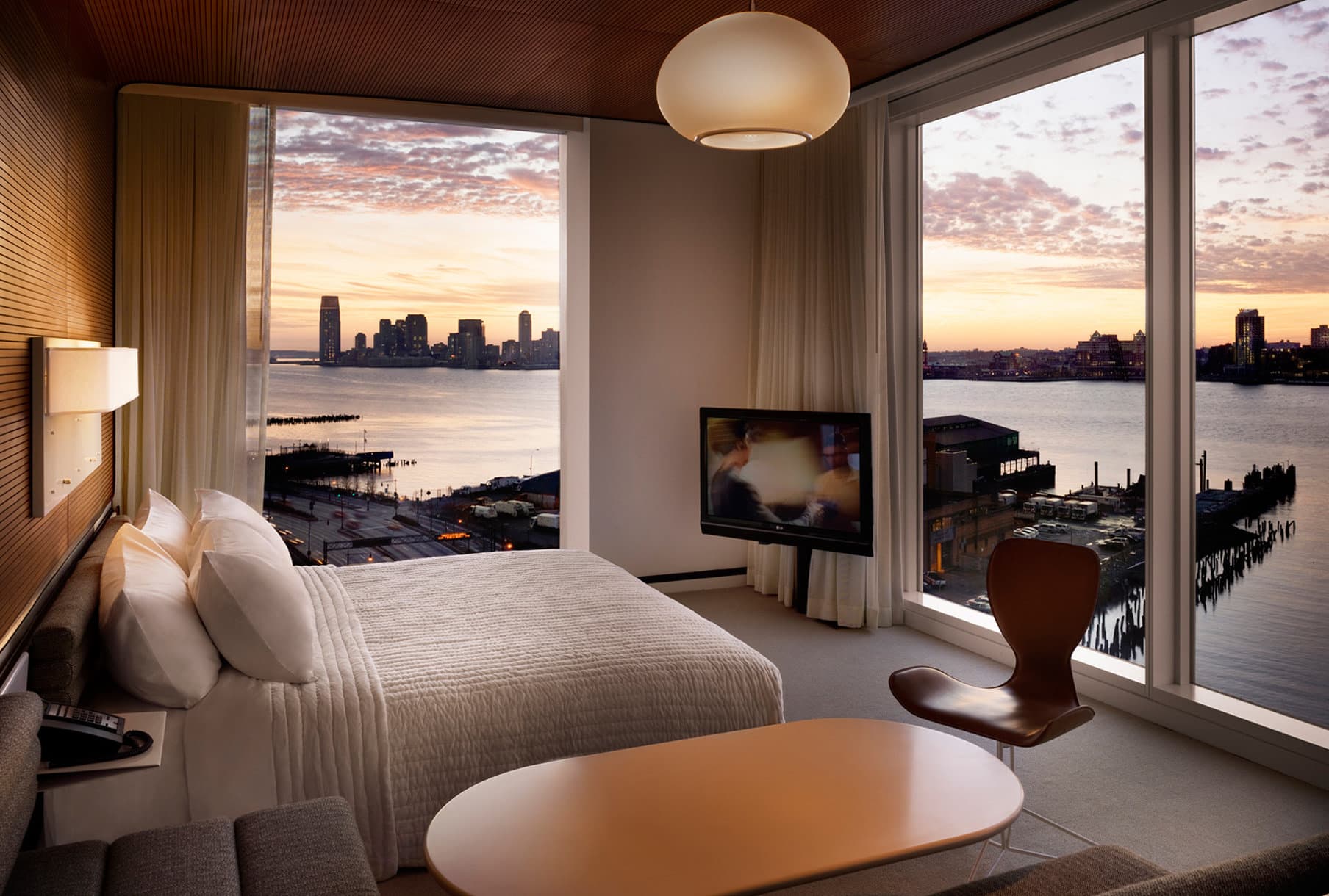 A guest in a white hooded robe sitting in a floor-to-ceiling window at The Standard High Line, overlooking the Hudson River at sunset.