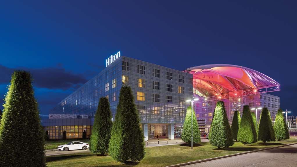 A towering 24-meter glass atrium filled with live palm trees and illuminated by soft evening lights at Hilton Munich Airport.