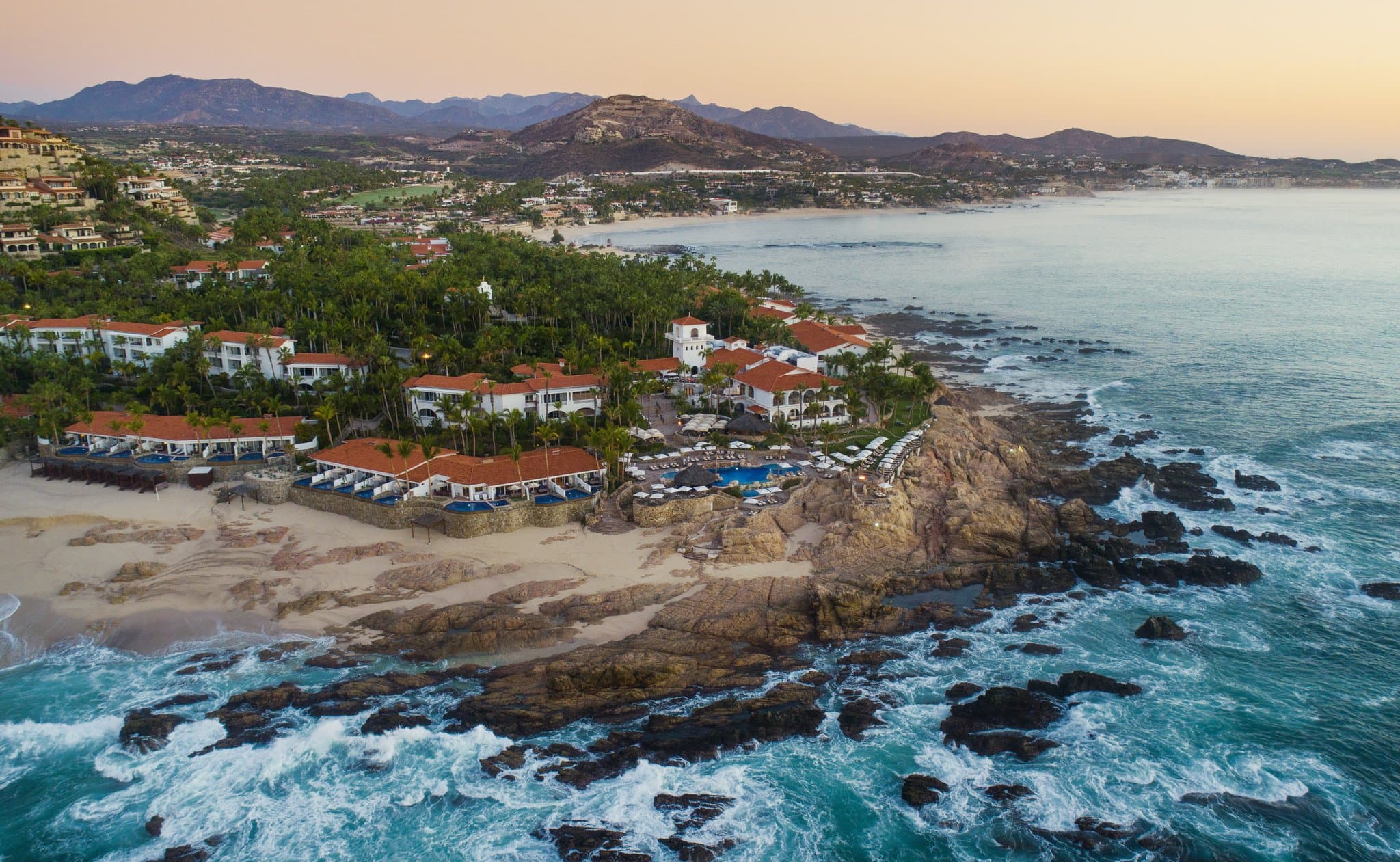 A dramatic view of the One&Only Palmilla's adults-only Vista Pool at sunset, framed by palm trees with the Sea of Cortez in the background.