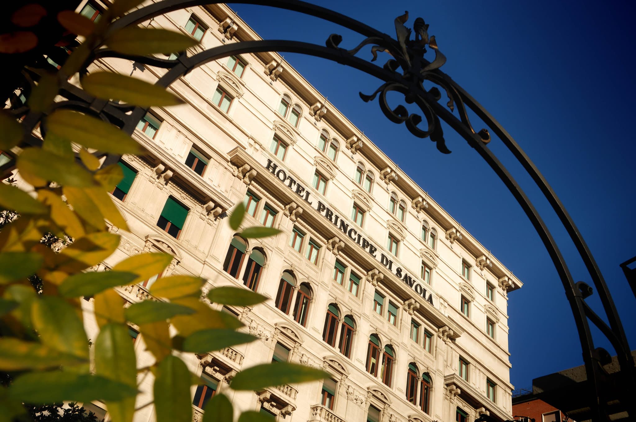 The illuminated neoclassical façade of Hotel Principe di Savoia at twilight, with luxury cars parked at the entrance.