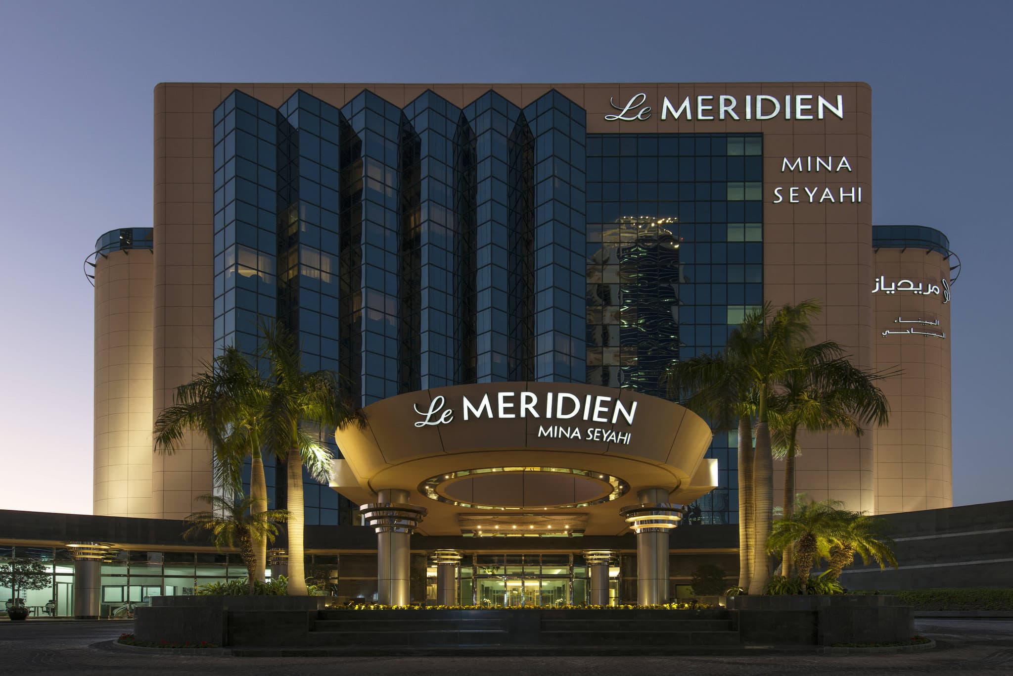 View of Le Méridien Mina Seyahi's Horizon Infinity Pool merging with the blue Arabian Gulf, framed by palm trees.