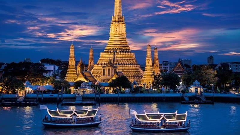 The Peninsula Bangkok's signature three-tiered swimming pool lined with palm trees, overlooking the Chao Phraya River and city skyline at dusk.