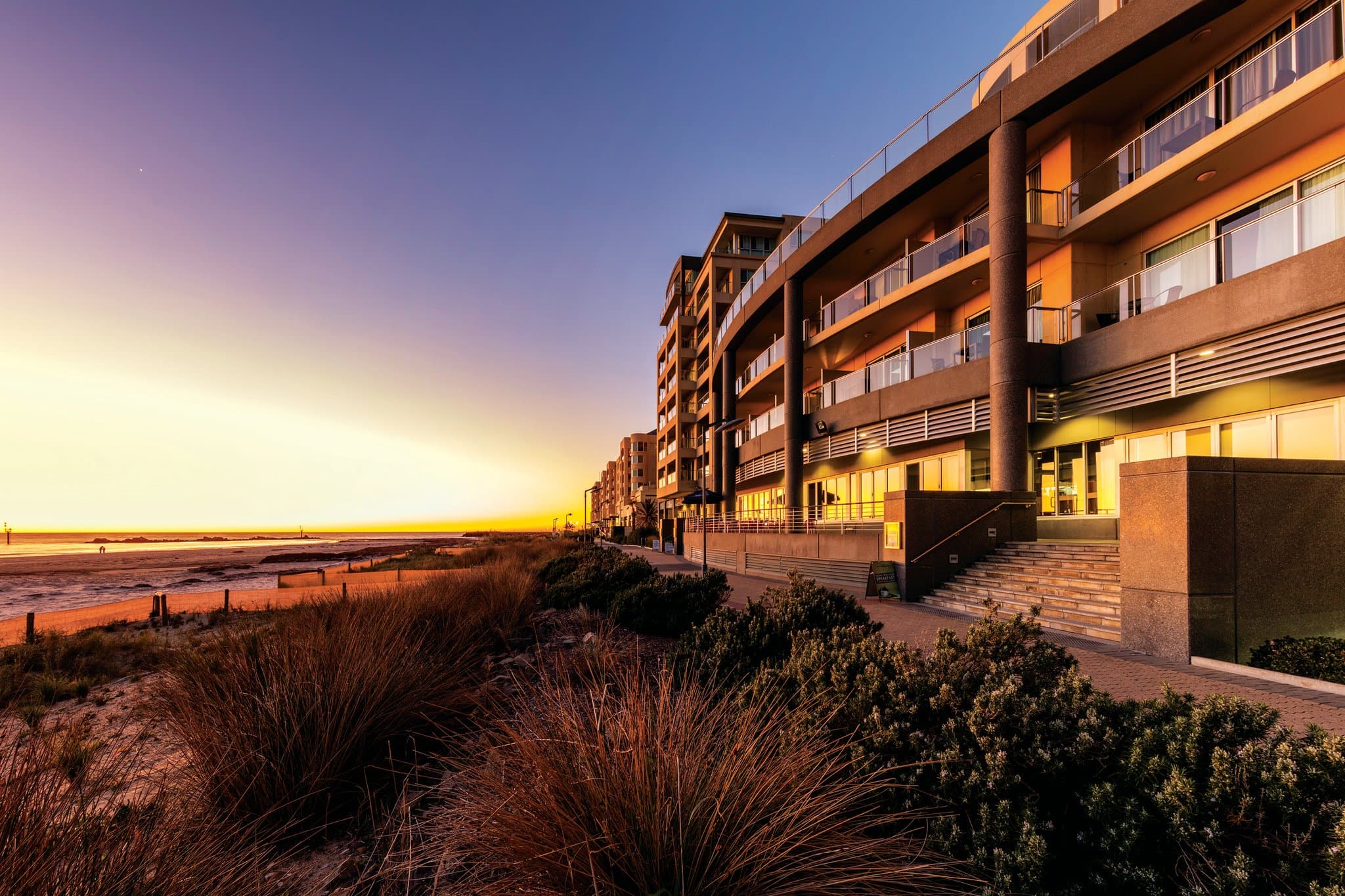View from Oaks Glenelg balcony at sunset, overlooking the Glenelg Jetty and the white facade of The Beachouse complex.