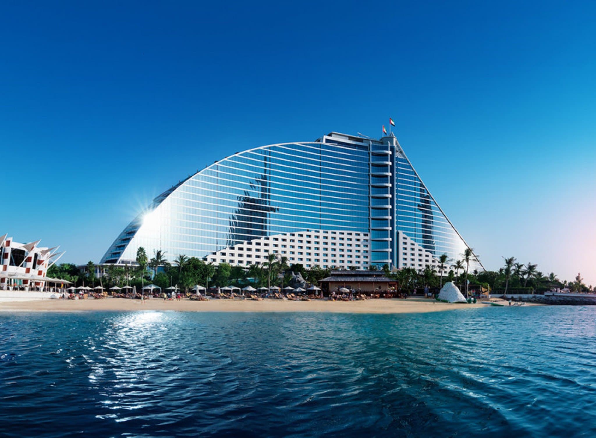 Jumeirah Beach Hotel's wave-shaped architecture framing the Burj Al Arab at sunset, with pools in the foreground.