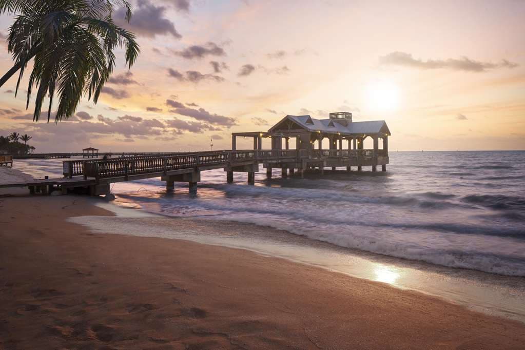 A couple walking on the private natural sand beach of The Reach Key West at sunset, with the iconic gazebo pier in the background.