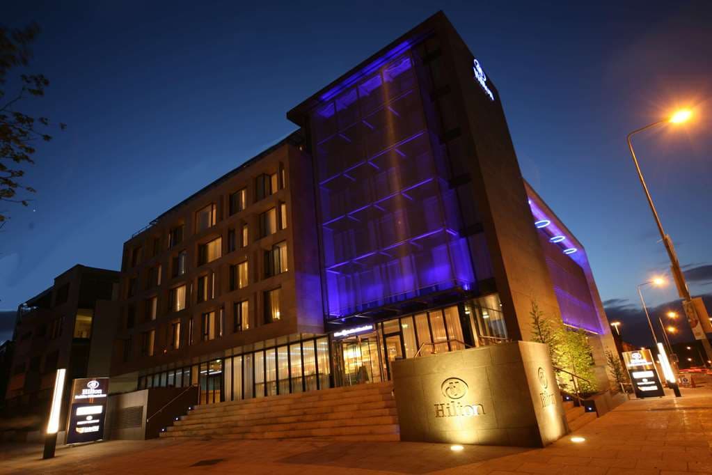 Hilton Dublin Kilmainham exterior at dusk with warm lighting, showcasing the glass façade opposite the historic Kilmainham Gaol walls.